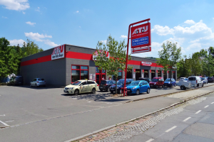 Ein Autohaus mit mehreren geparkten Autos vor einem Gebäude, umgeben von weiteren Gebäuden, Bäumen, Strommasten, Schildern und einer Straße unter einem bewölkten Himmel.