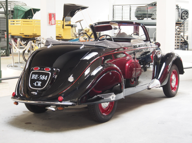 Ein schwarzer und roter Wagen in einer Garage geparkt, umgeben von anderen Fahrzeugen, Pfosten, Ketten und Gegenständen, mit einem Feuerlöscher an der Wand im Hintergrund.