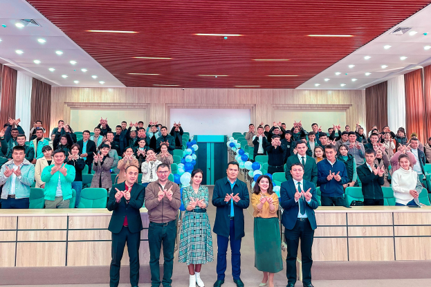 Eine Gruppe von Menschen steht vor einem Publikum, einige klatschen, in einem Raum mit Stühlen, Tischen, Ballons, Vorhängen und Deckenleuchten, bei der Eröffnungsfeier einer neuen Schule.