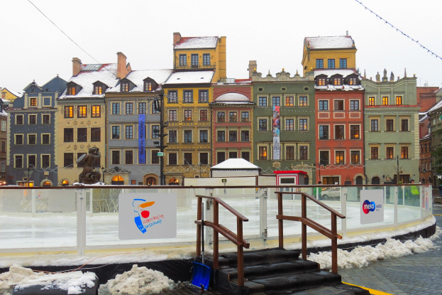 Eislaufbahn in einem Stadtplatz umgeben von Gebäuden, Treppen, Geländern, Informationsschildern, schneebedecktem Boden, Laternen, Bäumen und einem Himmel.