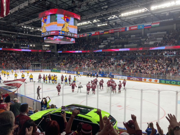 Hockey game in a crowded arena with spectators, rink fencing, banners, ceiling lights, a large screen, and hanging flags.