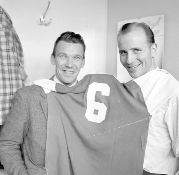 Zwei Männer in Schwarz-Weiß mit strahlendem Lächeln, die ein Fußballtrikot hochhalten, vor einer Wand mit einer Tafel und einem Bügel im Hintergrund.