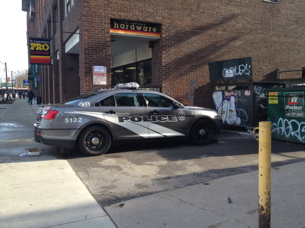 Polizeiauto auf der Straße vor einem Gebäude mit Fenstern und Namensschildern geparkt, mit Graffiti-bedeckten Mülltonnen rechts und einem Pfahl im Vordergrund, Bäume und einen klaren blauen Himmel im Hintergrund.