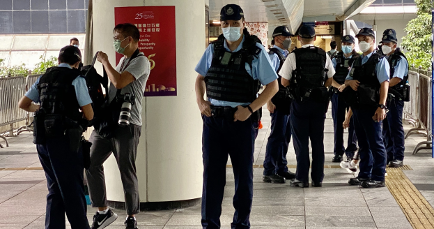 Eine Gruppe von Polizisten in Uniform, Mützen und Mund-Nasen-Schutz steht vor einem Gebäude, einige halten Kameras, mit einem Banner an einer Säule, Geländern, Pflanzen, Bäumen und Deckenleuchten im Hintergrund.