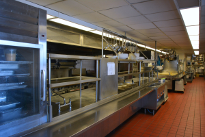 Kommerzielle Küche mit Edelstahl-Theke, hängenden Utensilien, einem Spülbecken, Wasserhähnen, Schränken und diversen Gegenständen, die in Regalen unter elektrischen Deckenleuchten angeordnet sind.