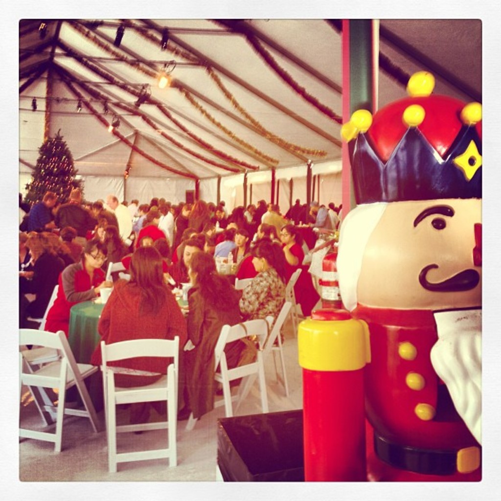 Eine Gruppe von Menschen feiert Weihnachten in einer Hütte, mit einem geschmückten Baum, weißen Stühlen, einem roten Spielzeug-Sicherheitswächter und einer schwarzen Krone.