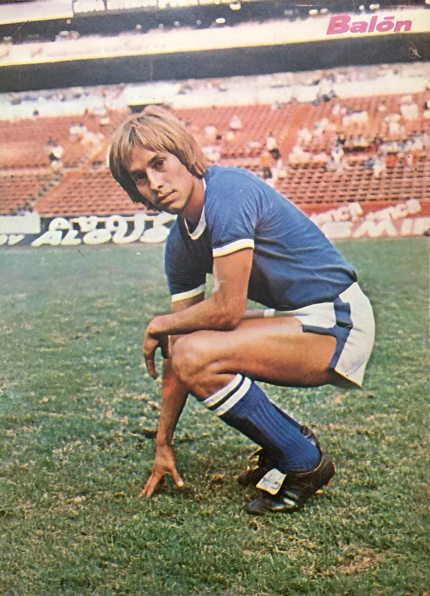 Ein Mann kniet auf einem Fußballfeld umgeben von Menschen mit Schildern, trägt Sportschuhe.