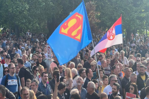 Eine große Gruppe von Menschen steht vor einer Menge, einige halten Fahnen und Schilder mit der Aufschrift "Serbischer Anti-Superman-Protest", mit Bäumen und Gras im Hintergrund.