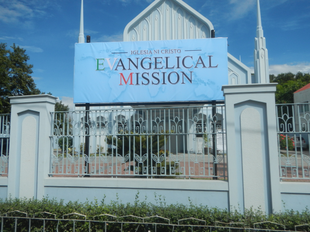 Eine Kirche mit einem Schild, auf dem "Evangelische Mission" steht, ist von einem Metallzaun, Pflanzen, Bäumen und einem Haus mit einem Dach umgeben, unter einem bewölkten Himmel.
