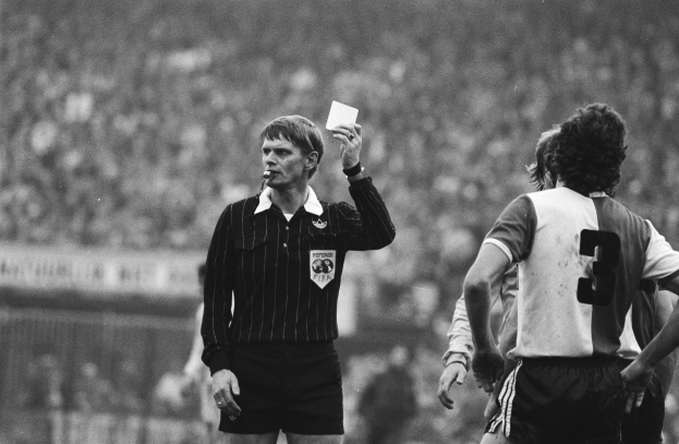 Schwarzes und weißes Bild eines Schiedsrichters, der eine rote Karte vor einer Menge von Menschen mit Fußballtrikots hält, mit einem Zaun im Hintergrund.