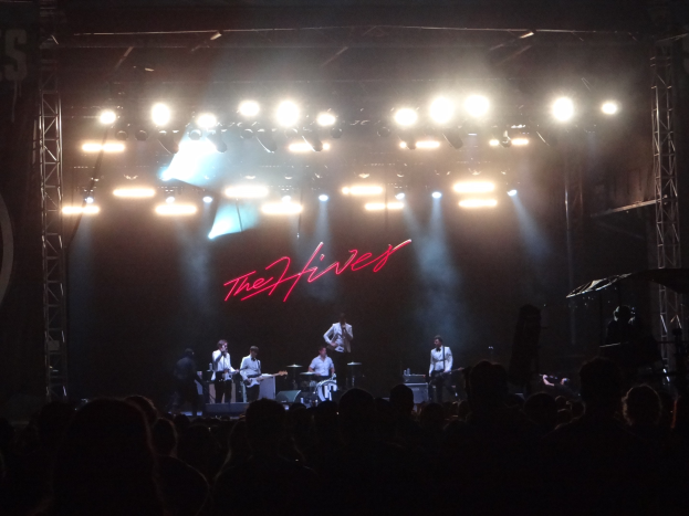 Eine Gruppe von Musikern performs auf der Bühne eines Konzerts, wobei ein Sänger ein Mikrofon hält, während das Publikum darunter zusieht.