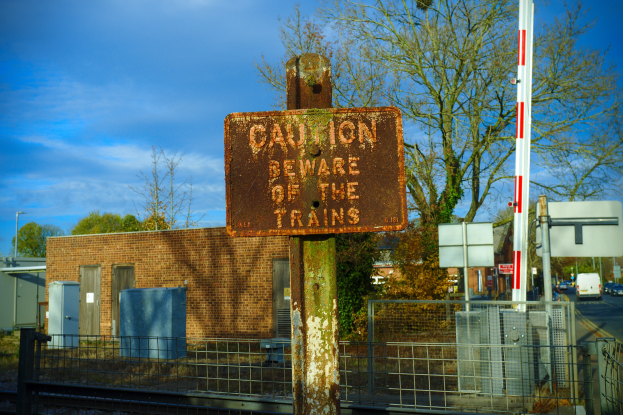 Warnschild an einem Bahnsteigzaun mit Bäumen, Strommasten, einem Gebäude, Containern, Fahrzeugen auf einer Straße und einem bewölkten Himmel.