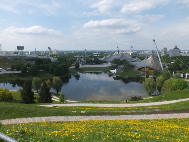 Blick auf das Olympiastadion von einem Hügel aus, mit einem Teich im Vordergrund, umgeben von Grün und gelben Blumen, Gebäuden und Pfählen im Hintergrund unter einem klaren blauen Himmel.
