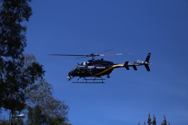 Polizeihubschrauber fliegt durch einen blauen Himmel mit Bäumen im Vordergrund und einem Lichtmast auf der linken Seite.