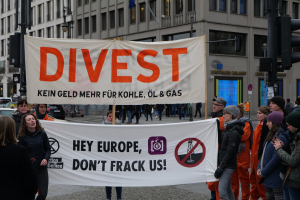 Eine Gruppe von Menschen hält ein Transparent mit der Aufschrift "Divest Hey Europe, Don't Frack Us" auf einer Straße mit Gebäuden, Straßeninfrastruktur, Fahrzeugen, Bäumen und einem klaren Himmel im Hintergrund.