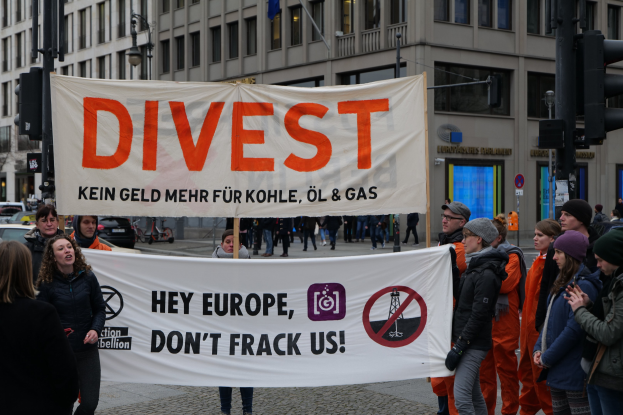 Eine Gruppe von Menschen hält ein Transparent mit der Aufschrift "Divest Hey Europe, Don't Frack Us" auf einer Straße mit Gebäuden, Straßeninfrastruktur, Fahrzeugen, Bäumen und einem klaren Himmel im Hintergrund.