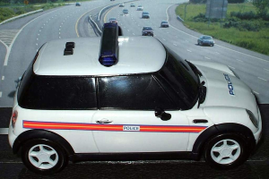 Weißer Polizeiwagen vor fahrendem Verkehr auf einer Straße mit Gras und Bäumen außerhalb der Stadt.