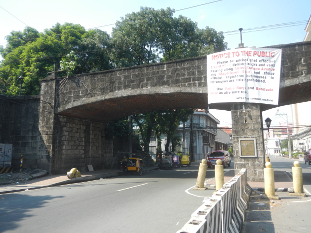 Öffentliche Ankündigungstafel auf einer Brücke über einer Straße mit Fahrzeugen, einem Geländer auf der rechten Seite, Gebäuden, Bäumen, Strommasten, Drähten und Himmel im Hintergrund.