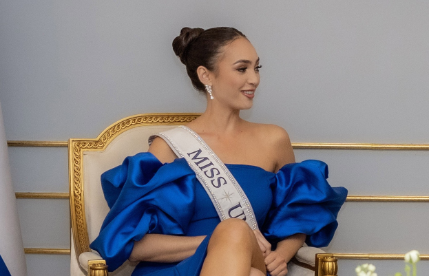 Frau in einem blauen Kleid mit einer "Miss Universe 2019"-Schärpe, die auf einem Stuhl sitzt und von Blumen umgeben ist und lächelt.