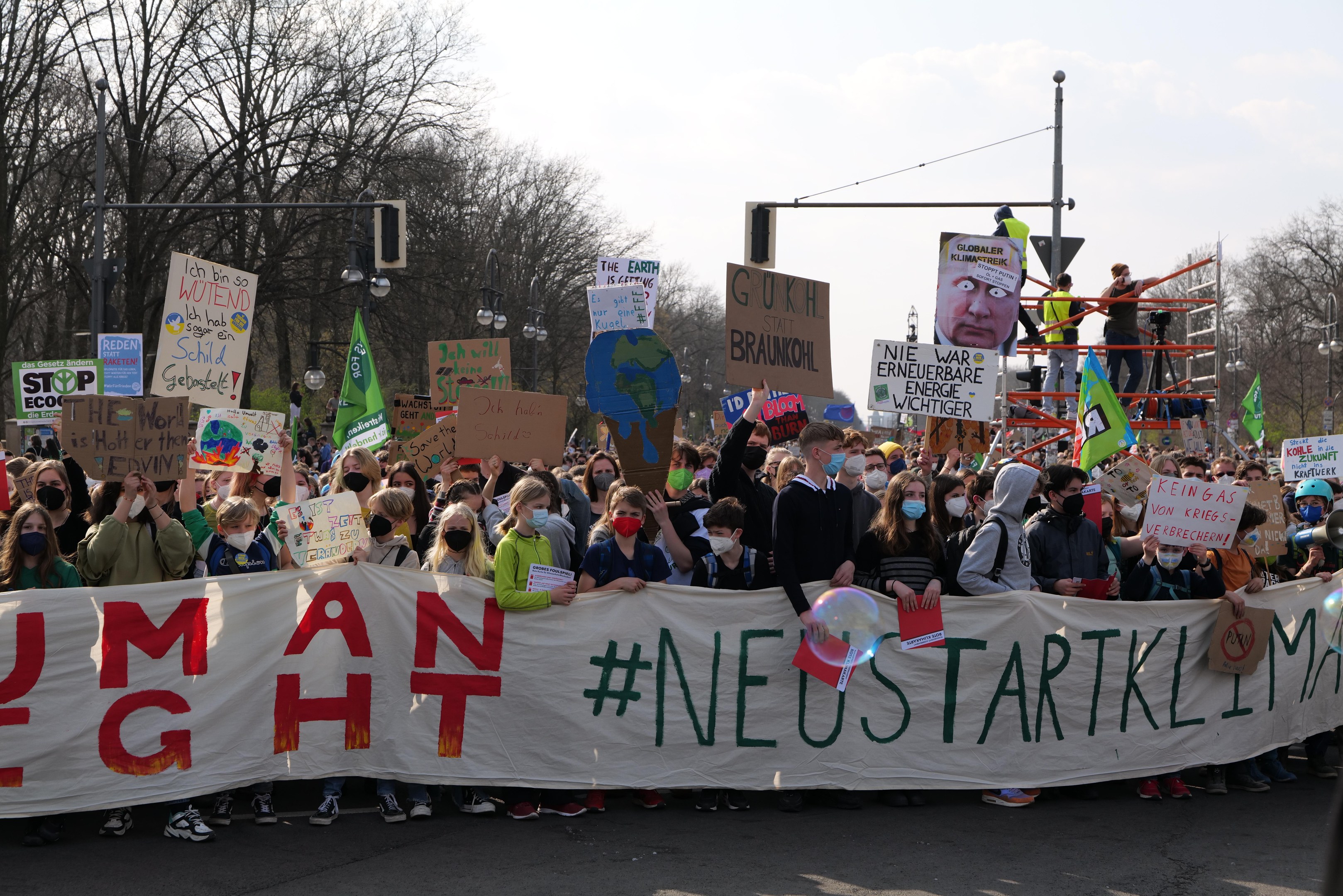 Eine große Gruppe von Menschen marschiert auf einer Straße, trägt ein 'Menschliche Rechte' -Schild und verschiedene Plakate, einige tragen Masken, mit Bäumen, Laternenmasten und einem klaren blauen Himmel im Hintergrund.