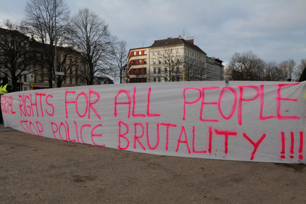 Eine Gruppe von Menschen, die auf dem Boden stehen und ein Banner halten, auf dem 'Rechte für alle Menschen Stoppt Polizeigewalt' steht, mit einem Straßenmast, einem Schild, Bäumen, Gebäuden mit Fenstern und einem bewölkten Himmel im Hintergrund.