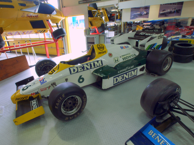 Eine Garage mit verschiedenen Rennwagen, Reifen, einem hölzernen Objekt, einem Geländer, einer Glastür, gerahmten Fotos an der Wand und Deckenbeleuchtung.
