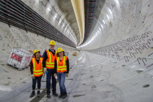Drei Personen in Sicherheitswesten und gelben Helmen stehen in einem Tunnel, mit einem weißen Kasten links und Rohren rechts.