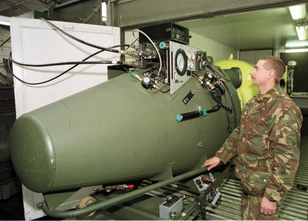 Ein Mann in militärischer Uniform steht neben einer großen grünen Bombe in einem unordentlichen Raum mit Maschinen, Drähten und beleuchteten Wänden, mit Text unten.