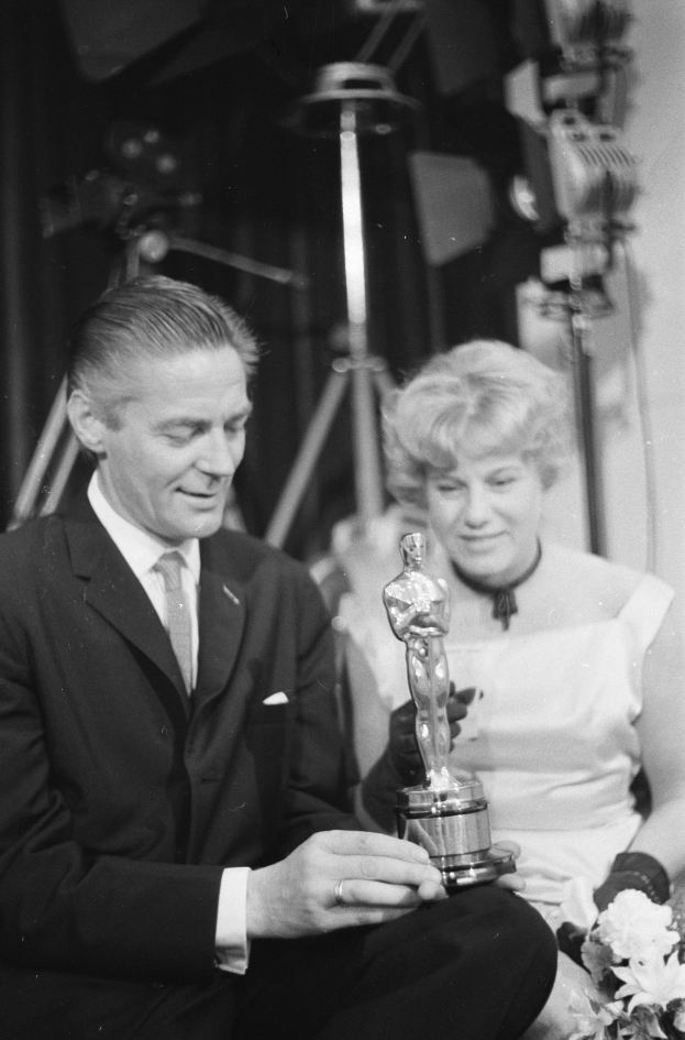 Ein Mann in einem schwarzen Blazer, weißem Hemd und Krawatte hält eine Oscar-Statue in der Hand und sitzt neben einer Frau, die einen Blumenstrauß hält, beide in eleganter Kleidung, mit Licht im Hintergrund, schwarz-weiß.