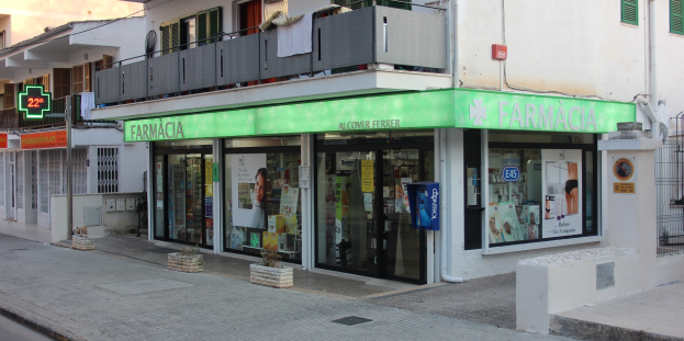 Eine Straßenansicht einer Apotheke mit benachbarten Gebäuden, sichtbarer Beschilderung, Strommasten, Stromkabeln, fahrenden Fahrzeugen, Topfpflanzen und einem klaren blauen Himmel.
