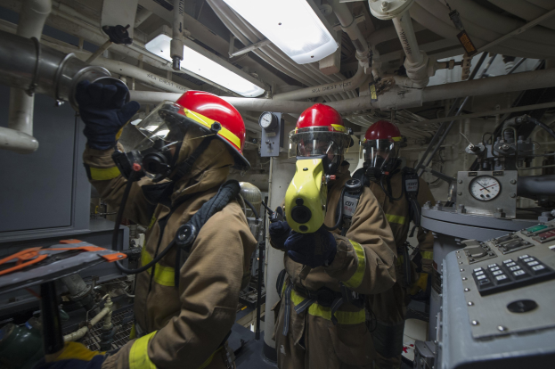 Drei Feuerwehrleute in Helmen und Gasmasken stehen vor einem Bedienpult mit einer Maschine rechts daneben und Rohren, Lampen und anderen Gegenständen im Hintergrund.