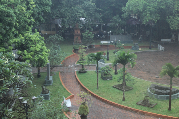 Ein grüner Park mit verschiedenen Bäumen, Pflanzen, Gras, Laternen, Bänken und Skulpturen mit einem Gebäude im Hintergrund.