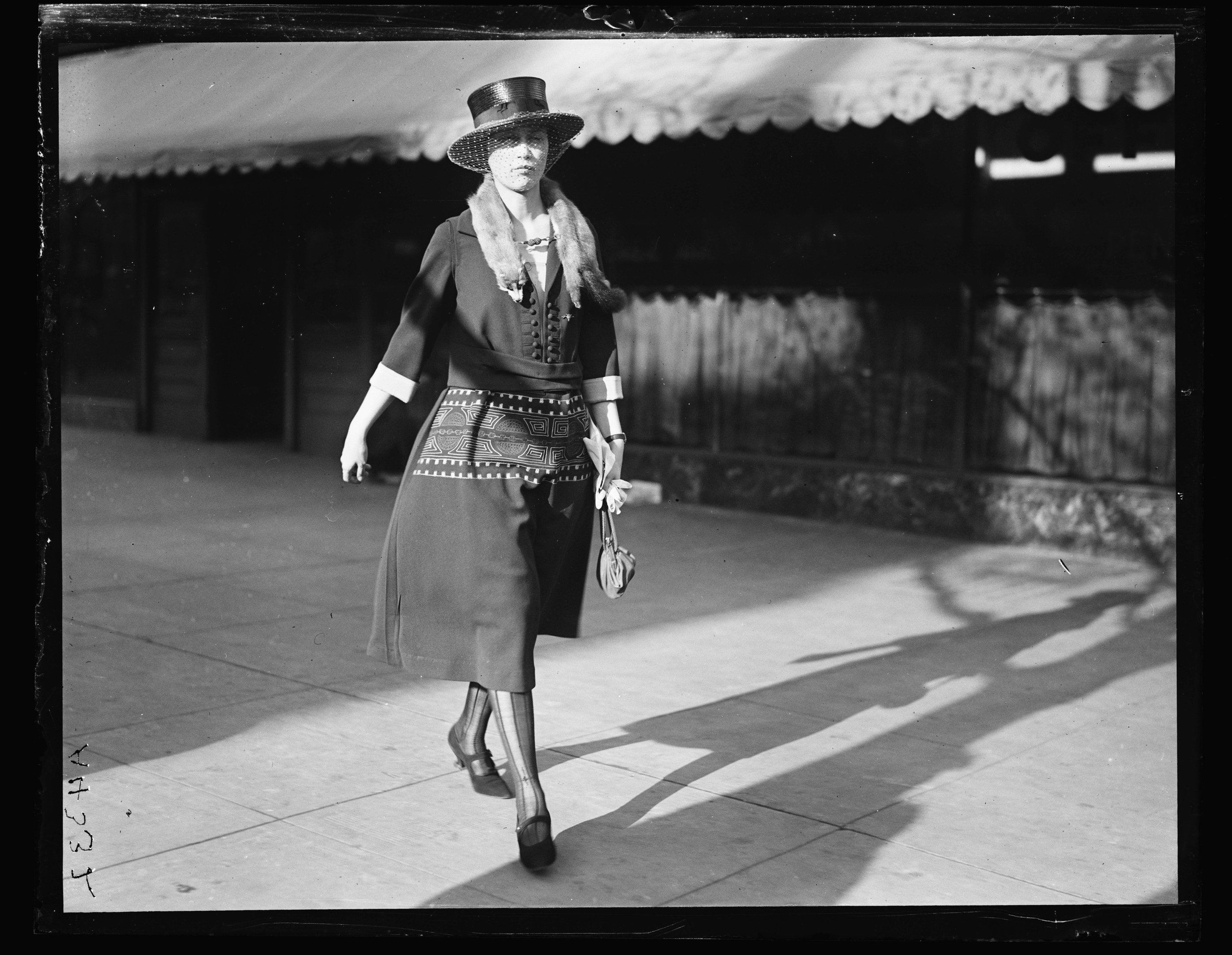 Eine stilvolle Frau in einem Kleid und Hut geht die Straße entlang in Schwarz-Weiß, hält eine Handtasche und trägt hohe Absätze.
