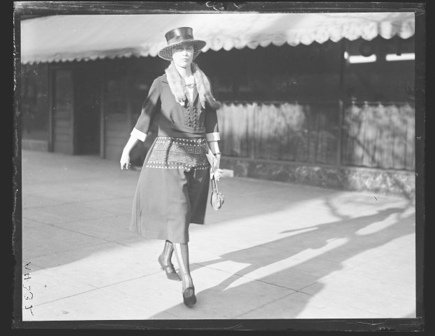 Eine stilvolle Frau in einem Kleid und Hut geht die Straße entlang in Schwarz-Weiß, hält eine Handtasche und trägt hohe Absätze.