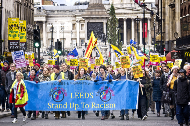 Eine Gruppe von Menschen marschiert mit einem 'Steh Auf gegen Rassismus'-Schild die Straße entlang, mit Gebäuden, Bäumen, Laternen, Verkehrsampeln und einer Statue im Hintergrund bei bewölktem Himmel.