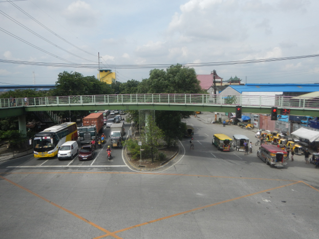 Verkehrsreiche Innenstadtstraße mit Autos, Bussen, Fußgängern, einer Brücke, Ampeln, Gebäuden, Bäumen und Strommasten.