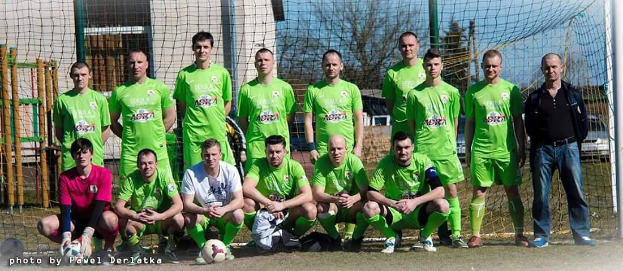 Eine Gruppe von Männer in grünen T-Shirts steht auf einem Fussballfeld, einige sind in Hockstellung, mit einem Ball und einem Torpfosten, umgeben von Bäumen, Geb&aumluden und Fahrzeugen.