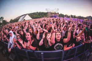Eine große Menschenmenge auf einem Musikfestival mit erhobenen Händen vor einem Geländer, mit Bäumen, Gebäuden und einem klaren blauen Himmel im Hintergrund.