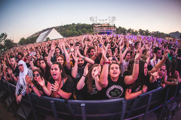 Eine große Menschenmenge auf einem Musikfestival mit erhobenen Händen vor einem Geländer, mit Bäumen, Gebäuden und einem klaren blauen Himmel im Hintergrund.