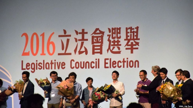 Gruppe von Menschen auf einer Bühne mit Blumenstr├Ąu├če, mit audienceheads visible below and a screen displaying text and a logo for the 2016 legislative council election in the background.