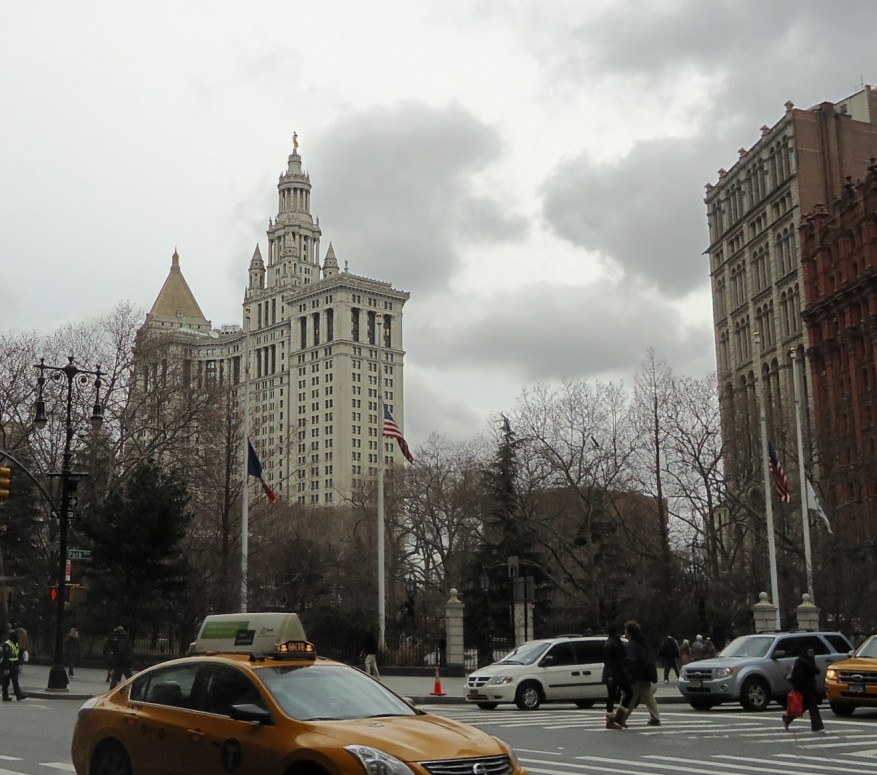Gelbes Taxi fährt auf einer Stadtstraße mit hohen Gebäuden, Fußgängern auf dem Gehweg, Verkehrskegeln, Laternenmasten, Fahnenmasten, Bäumen und einem bewölkten Himmel.