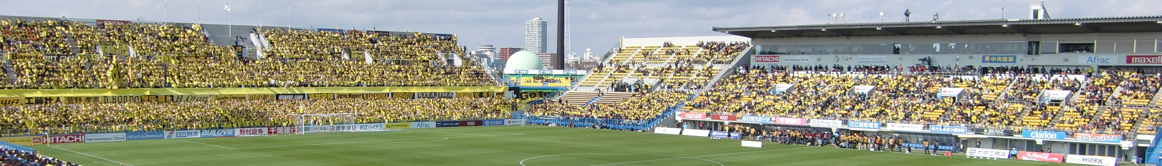 Großes Stadion voller Zuschauer bei einem Fußballspiel auf saftigem Grün, mit Werbetafeln, Gebäuden, Pfählen und einem klaren blauen Himmel im Hintergrund.