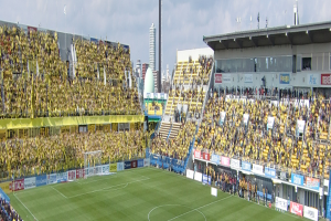 Großes Stadion voller Zuschauer bei einem Fußballspiel auf saftigem Grün, mit Werbetafeln, Gebäuden, Pfählen und einem klaren blauen Himmel im Hintergrund.