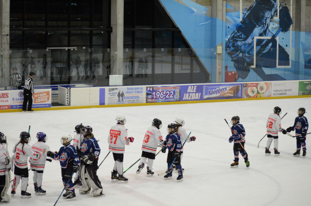 Gruppe junger Menschen, die Eis Hockey auf einer Indoor-Eisbahn spielen, mit Helmen und Hockey-Schlägern, sowie einer Person auf der linken Seite und Bannern, Säulen und Glaswänden im Hintergrund.