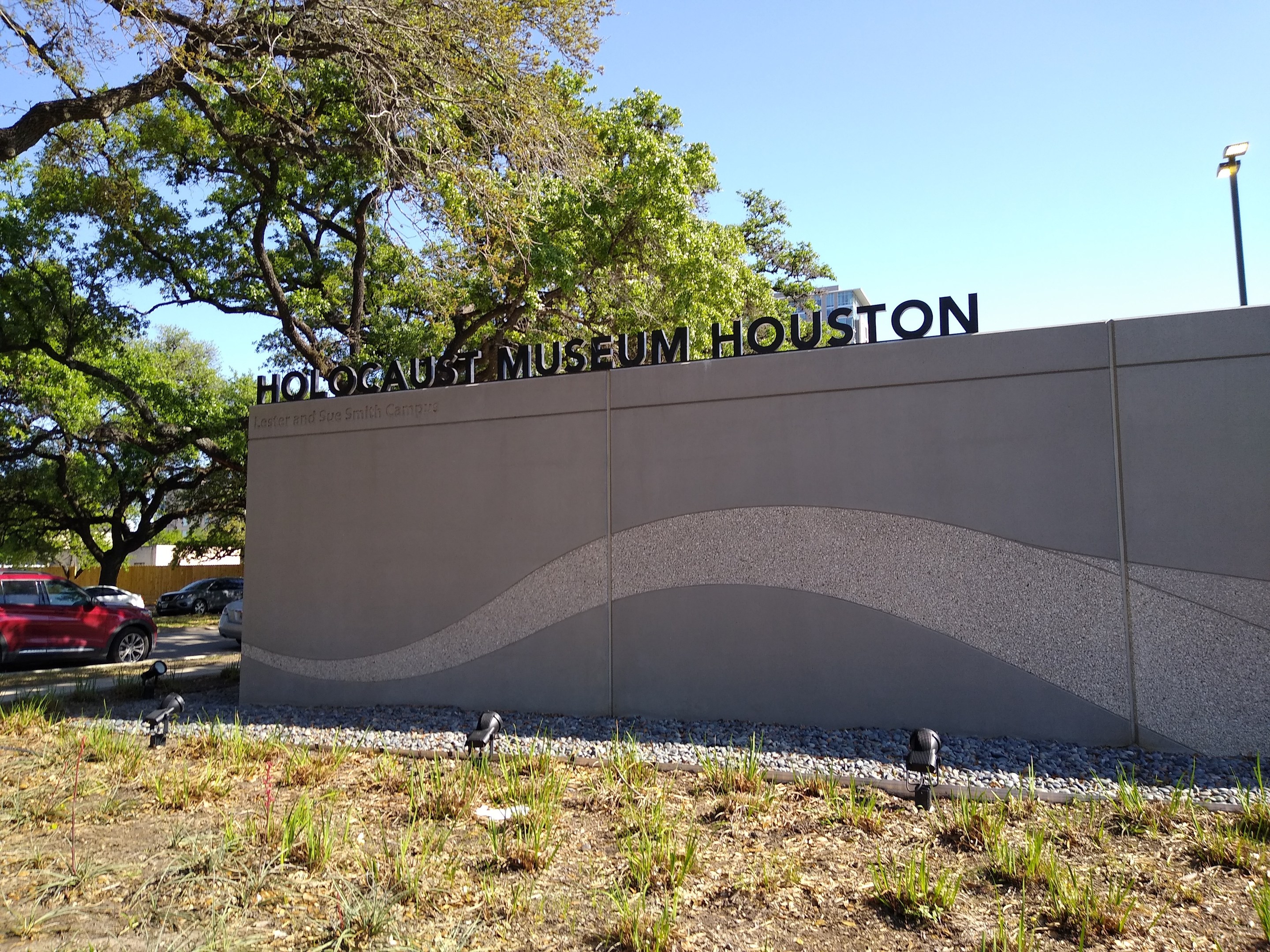 Außenansicht des Houston Holocaust Museums mit eingravierter Textwand, Gedenksteine, Grünfläche, Straße mit Fahrzeugen, einer Straßenlaterne und einem bewölkten Himmel.