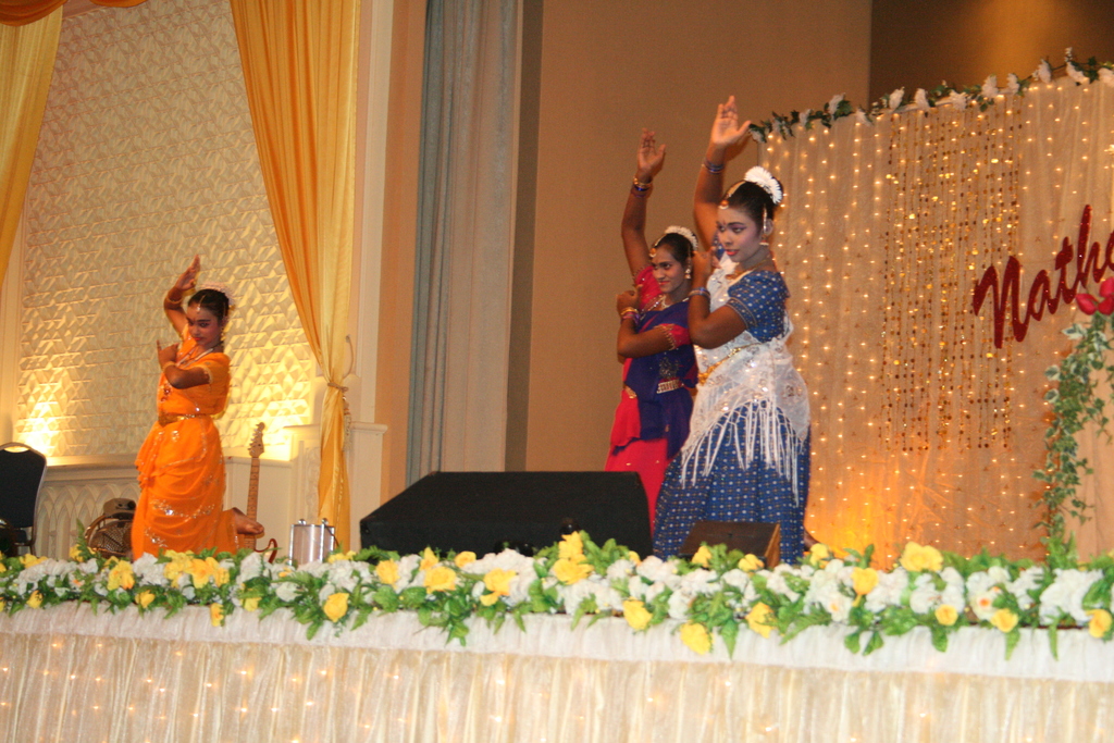 Eine Bühne mit weißen und gelben Blumen, drei tanzende Frauen, ein schwarzes Objekt, ein cremefarbener Vorhang, Lichter, eine braune Wand und andere Objekte im Hintergrund.