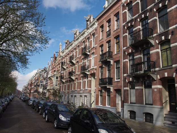 Eine Reihe geparkter Autos entlang einer von Bäumen gesäumten Straße in Amsterdam, mit Gebäuden, die Fenster, Geländer und Treppen aufweisen.