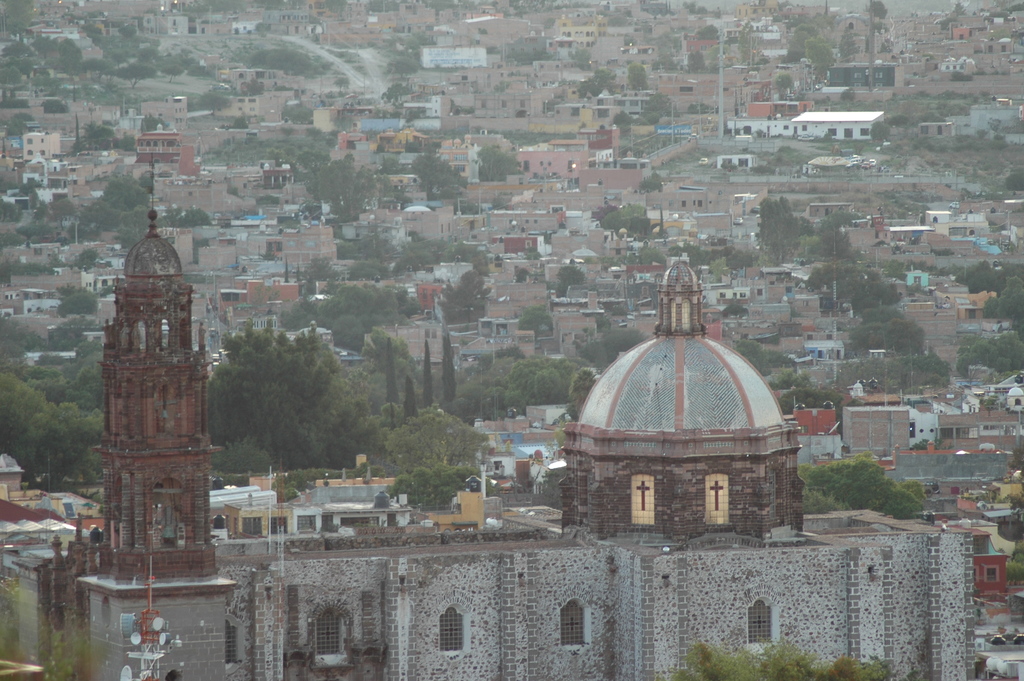 Luftaufnahme einer Stadt mit Bäumen, Gebäuden, Straßen, Masten und einer Kirche im Vordergrund.