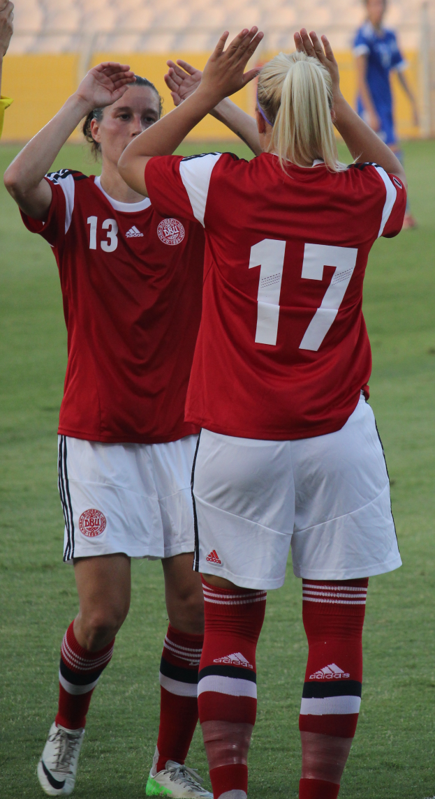 Zwei Frauen in roten T-Shirts und weißen Shorts stehen auf einem Fuballfeld und klatschen in die Hände, mit einer Person in einem blauen Kleid, einer gelben Tafel und einigen Stühlen im Hintergrund.