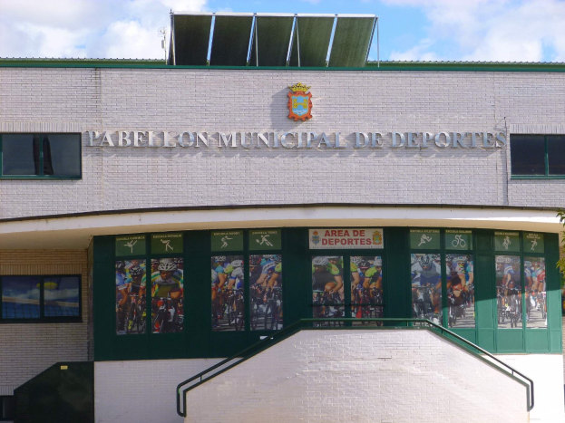 Ein Gebäude mit einer Tafel, die "Pabellón Municipal de Deportes" trägt, mit Fenstern, einer Treppe, einem Metallzaun, Pflanzen und Bäumen unter einem bewölkten Himmel.